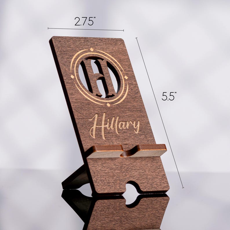 May include: A wooden phone stand, 5.5 inches tall and 2.75 inches wide, with a personalized design. The stand features a cutout initial within a circular emblem and the name "Hillary" in script. The wood has a dark brown finish.