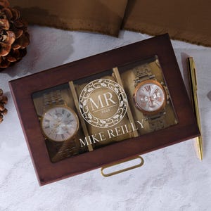 May include: A wooden watch box with a clear top, displaying two gold-toned watches and a decorative center. The center features the initials "MR" and the name "Mike Reilly" in white. A gold pen and a small leather wallet are also visible.