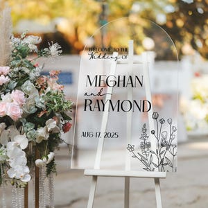 May include: Clear acrylic wedding sign on a white easel. The sign reads "Welcome to the Wedding of Meghan and Raymond, Aug 17, 2025" in black script. Floral line art decorates the bottom right corner. A floral arrangement is to the left.