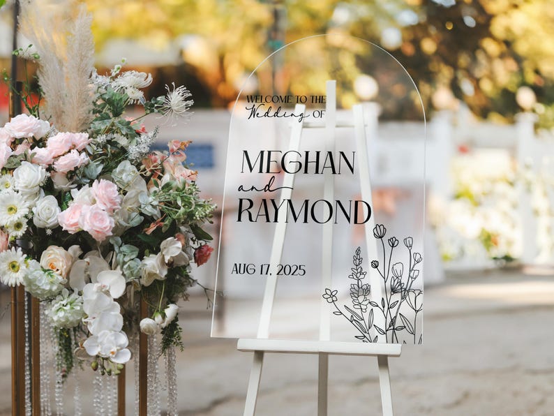 May include: Clear acrylic wedding sign on a white easel. The sign reads "Welcome to the Wedding of Meghan and Raymond, Aug 17, 2025" in black script. Floral line art decorates the bottom right corner. A floral arrangement is to the left.
