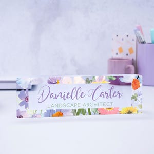 May include: A clear acrylic desk nameplate with a floral design featuring purple, pink, yellow, and green flowers. The nameplate reads "Danielle Carter Landscape Architect".