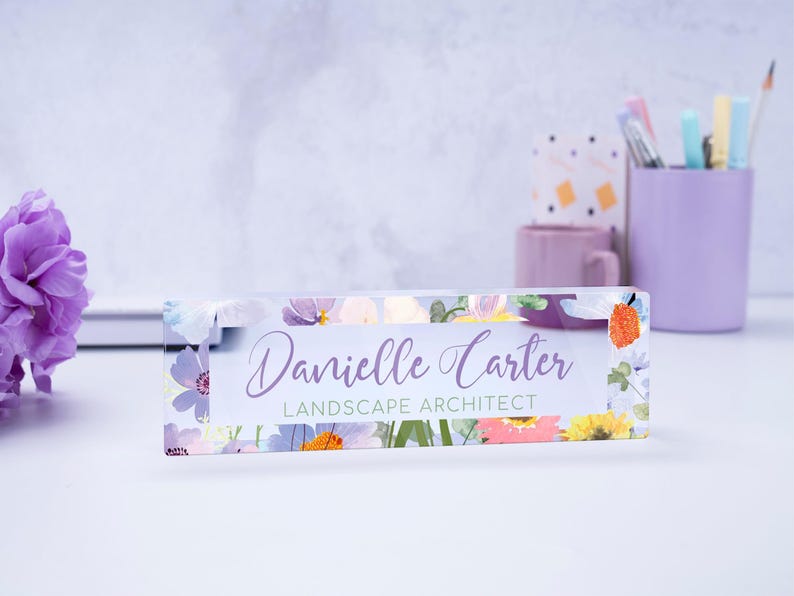 May include: A clear acrylic desk nameplate with a floral design featuring purple, pink, yellow, and green flowers. The nameplate reads "Danielle Carter Landscape Architect".