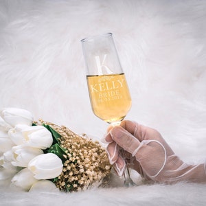 Może przedstawiać: Kieliszek do szampana z wygrawerowanym imieniem "KELLY", słowem "BRIDE" i datą "06.03.2023" na szkle. Kieliszek jest wypełniony złotym płynem i trzymany w pobliżu bukietu białych tulipan&oacute;w i suszonych kwiat&oacute;w.