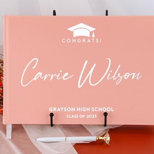 May include: A pink graduation guest book with a white graduation cap and the text "Congrats!" on the front. The name "Carrie Wilson" is written in white cursive script below the cap. The text "Grayson High School Class of 2025" is printed in white at the bottom of the book.