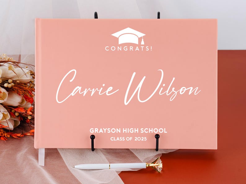 May include: A pink graduation guest book with a white graduation cap and the text "Congrats!" on the front. The name "Carrie Wilson" is written in white cursive script below the cap. The text "Grayson High School Class of 2025" is printed in white at the bottom of the book.