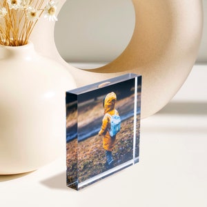 May include: A clear acrylic photo block displaying a color image of a child in a yellow coat and blue backpack. The block is rectangular and stands upright, showcasing the photograph. A white vase with dried flowers and a beige decorative object are in the background.