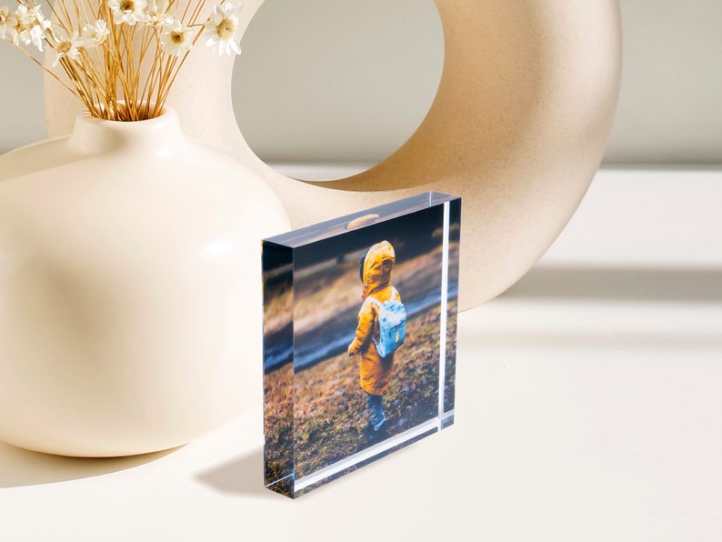 May include: A clear acrylic photo block displaying a color image of a child in a yellow coat and blue backpack. The block is rectangular and stands upright, showcasing the photograph. A white vase with dried flowers and a beige decorative object are in the background.