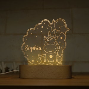 May include: A glowing unicorn night light with the name "Sophia" etched into the acrylic. The design features a cartoon unicorn, stars, and balloons, set on a wooden base. The light emits a warm, inviting glow.