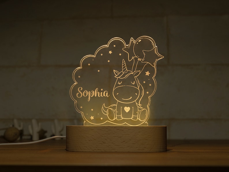 May include: A glowing unicorn night light with the name "Sophia" etched into the acrylic. The design features a cartoon unicorn, stars, and balloons, set on a wooden base. The light emits a warm, inviting glow.