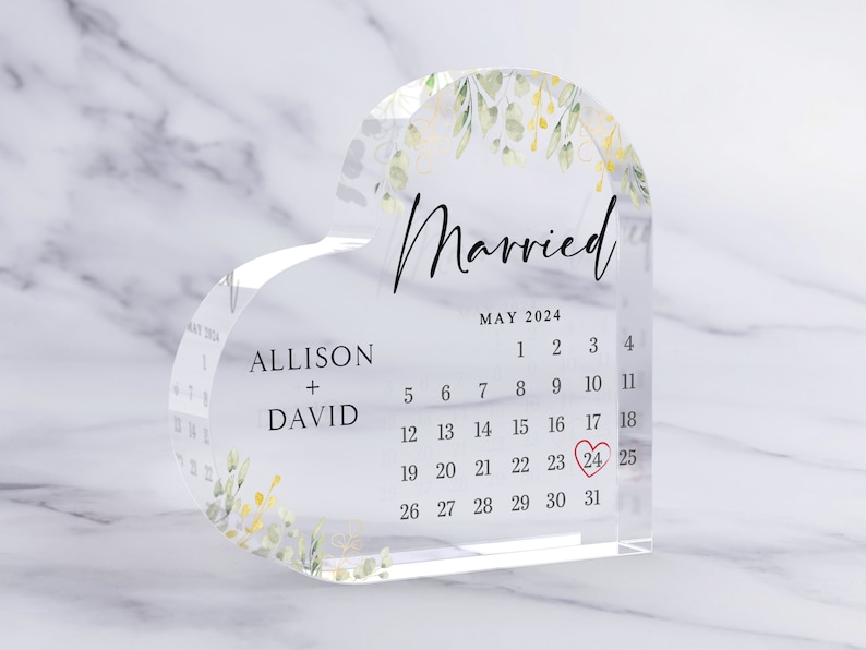 Puede incluir: Un calendario acr&iacute;lico transparente en forma de coraz&oacute;n con un dise&ntilde;o floral y la palabra "Casado" en la parte superior. El calendario es para mayo de 2024 y tiene un coraz&oacute;n rojo marcando el 24.