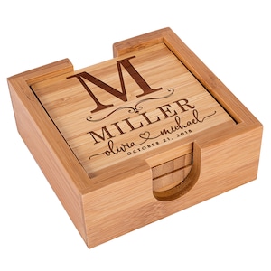 May include: A set of bamboo coasters in a matching holder. The top coaster is engraved with the letter "M", the last name "Miller", and the names "Olivia & Michael" with the date "October 21, 2018".
