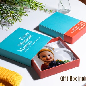 May include: A heart-shaped photo frame inside a red box with a teal lid. The lid reads "Every Moment Matters, Frame it Beautifully." The box also includes the text "Unveil Your Unforgettable Moments." A yellow beanie is visible.