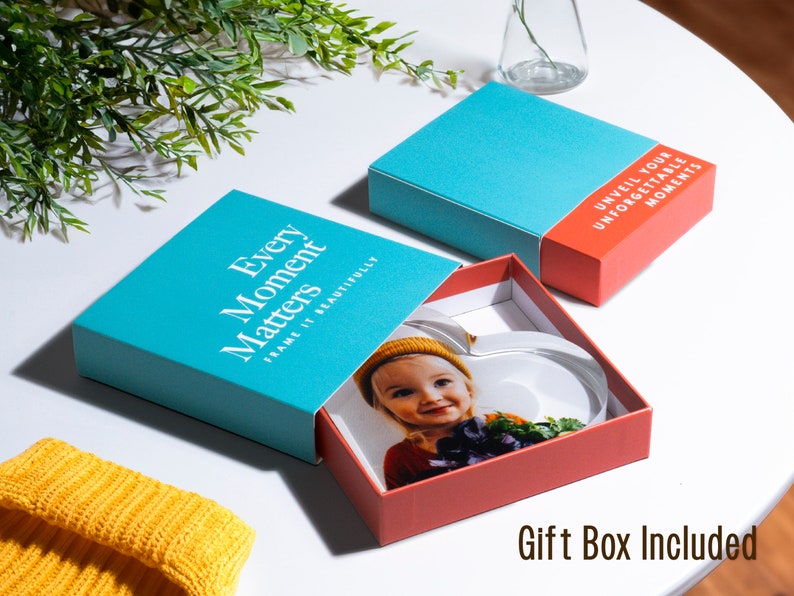 May include: A heart-shaped photo frame inside a red box with a teal lid. The lid reads "Every Moment Matters, Frame it Beautifully." The box also includes the text "Unveil Your Unforgettable Moments." A yellow beanie is visible.