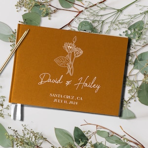 Op de afbeelding: Een mosterdgeel gastenboek met witte lintjes. De omslag heeft een lijntekening van een bloemenboeket en de tekst "David & Hailey" met de datum "July 11, 2024" en de locatie "Santa Cruz, CA".