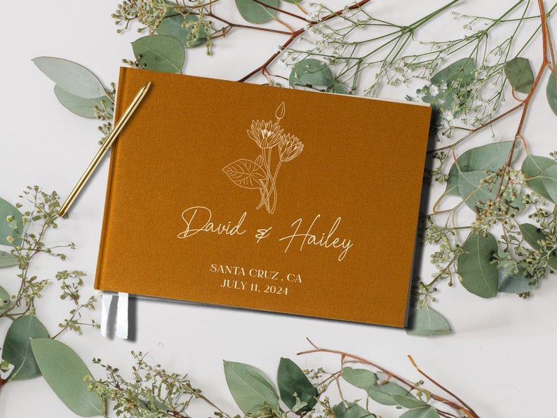 Op de afbeelding: Een mosterdgeel gastenboek met witte lintjes. De omslag heeft een lijntekening van een bloemenboeket en de tekst "David & Hailey" met de datum "July 11, 2024" en de locatie "Santa Cruz, CA".