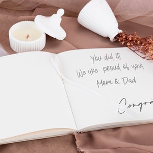 May include: A white book with a message written on the right page that says "You did it! We are proud of you! Mom & Dad Congrats!" The book is open and lying on a brown surface with a white candle and a white vase with dried flowers.