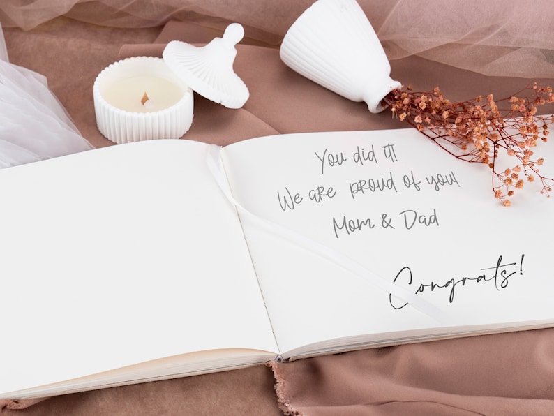 May include: A white book with a message written on the right page that says "You did it! We are proud of you! Mom & Dad Congrats!" The book is open and lying on a brown surface with a white candle and a white vase with dried flowers.