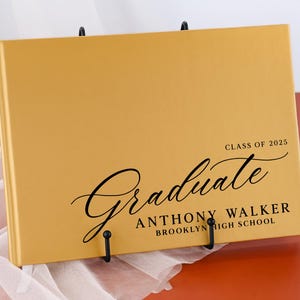 May include: A gold-colored graduation guest book with the text "Graduate" in a black cursive font. The book also includes the text "Class of 2025", "Anthony Walker", and "Brooklyn High School".