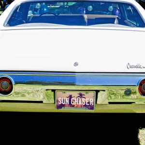 May include: A white vintage car with a license plate that reads "SUN CHASER". The license plate has a sunset and palm trees design.