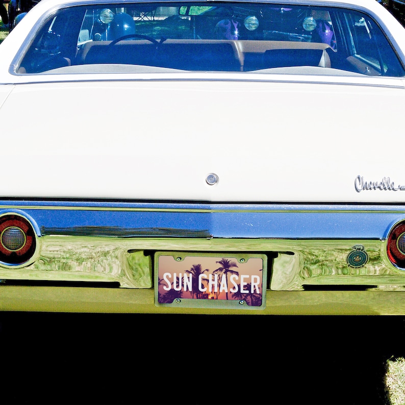 May include: A white vintage car with a license plate that reads "SUN CHASER". The license plate has a sunset and palm trees design.