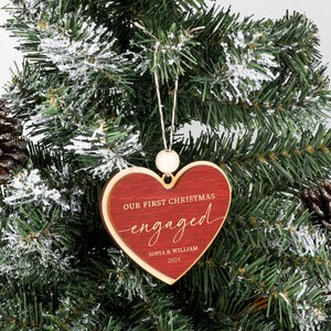 May include: Red wooden heart-shaped Christmas ornament with the text "Our First Christmas engaged" and the names "Sophia & William" and the year "2023".