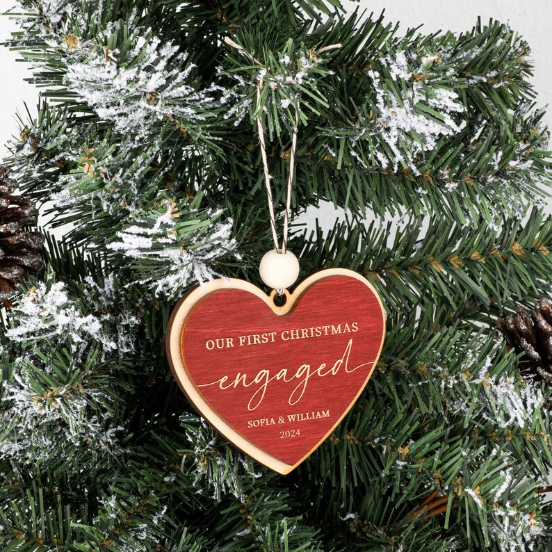 May include: Red wooden heart-shaped Christmas ornament with the text "Our First Christmas engaged" and the names "Sophia & William" and the year "2023".