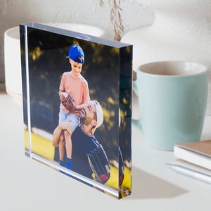May include: A clear acrylic photo block displaying a color photograph of a father and son. The son, wearing a blue baseball cap and a pink shirt, is sitting on his father's shoulders. The father is wearing a white cap and a dark blue shirt.