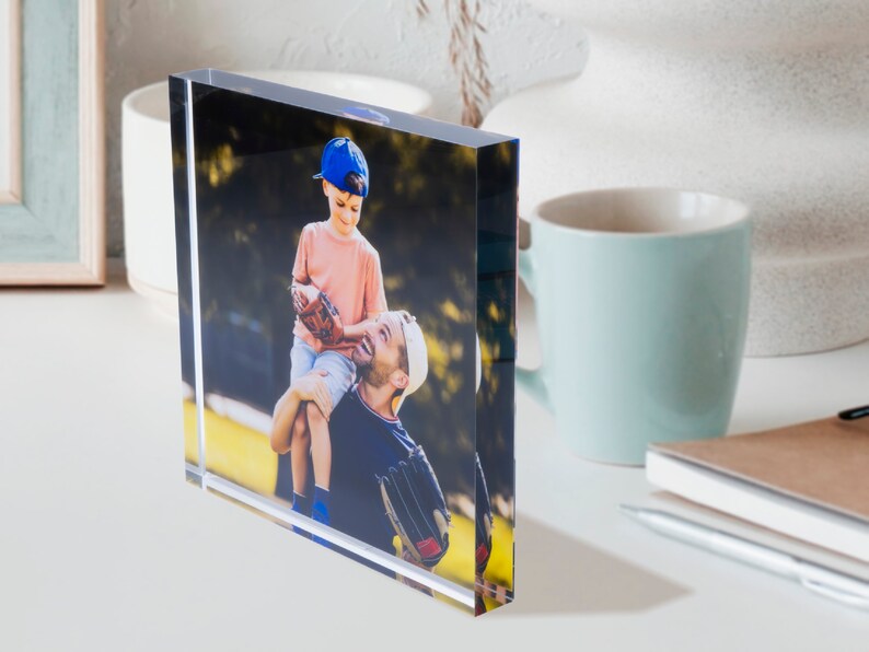 May include: A clear acrylic photo block displaying a color photograph of a father and son. The son, wearing a blue baseball cap and a pink shirt, is sitting on his father's shoulders. The father is wearing a white cap and a dark blue shirt.