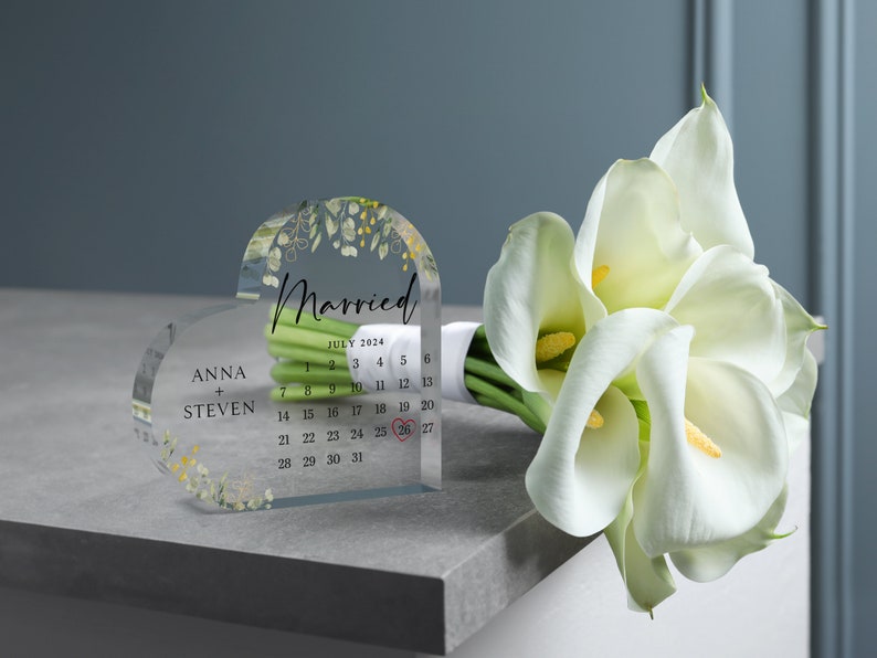 Puede incluir: Un recuerdo de boda de acr&iacute;lico transparente en forma de coraz&oacute;n con un dise&ntilde;o floral y la palabra "Casado" en &eacute;l. El recuerdo tambi&eacute;n incluye un calendario con la fecha "26 de julio de 2024" resaltada en rojo. Los nombres "Anna + Steven" est&aacute;n impresos en el recuerdo.