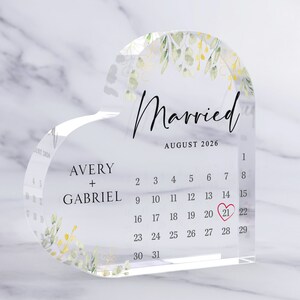 Puede incluir: Un calendario acr&iacute;lico transparente en forma de coraz&oacute;n con un dise&ntilde;o floral y el texto "Casado" y "Agosto 2026". La fecha "21" est&aacute; resaltada en rojo.