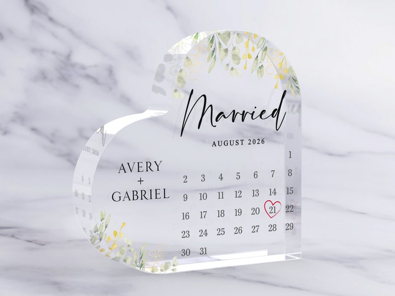 Puede incluir: Un calendario acr&iacute;lico transparente en forma de coraz&oacute;n con un dise&ntilde;o floral y el texto "Casado" y "Agosto 2026". La fecha "21" est&aacute; resaltada en rojo.