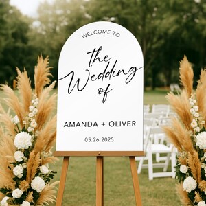 May include: A white wedding welcome sign on a wooden easel, with the text "Welcome to the Wedding of Amanda + Oliver 05.26.2025". The sign is flanked by floral arrangements with pampas grass and white flowers.