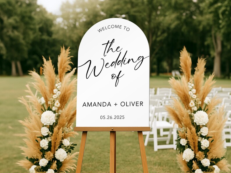 May include: A white wedding welcome sign on a wooden easel, with the text "Welcome to the Wedding of Amanda + Oliver 05.26.2025". The sign is flanked by floral arrangements with pampas grass and white flowers.