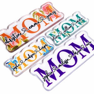 May include: Four acrylic plaques with the word "MOM" in large, colorful letters. Each plaque has a floral design and a personalized message with a heart and names, such as "Christine & Emma" and "Sophia & Melissa & Brian & Ava".