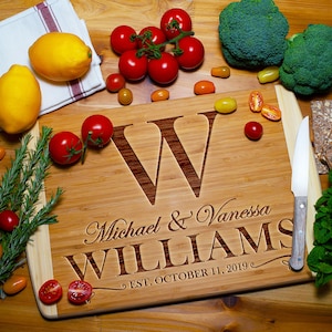 Może przedstawiać: Drewniana deska do krojenia z wygrawerowaną literą "W" oraz imionami "Michael & Vanessa WILLIAMS" z datą "EST. OCTOBER 11, 2019". Świeże produkty, w tym pomidory, brokuły i cytryny, otaczają deskę.