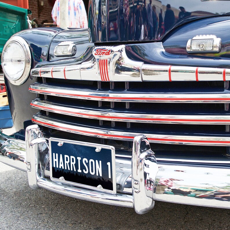 May include: A vintage blue Ford car with chrome accents and a red and white grille. The car has a license plate that reads "HARRISON 1".