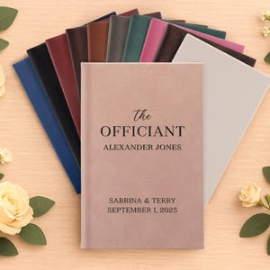 May include: A collection of personalized wedding officiant books in various colors, including pink, blue, and brown. The cover of the featured book is pink and reads "The Officiant" with a name and wedding date. Floral accents surround the books.