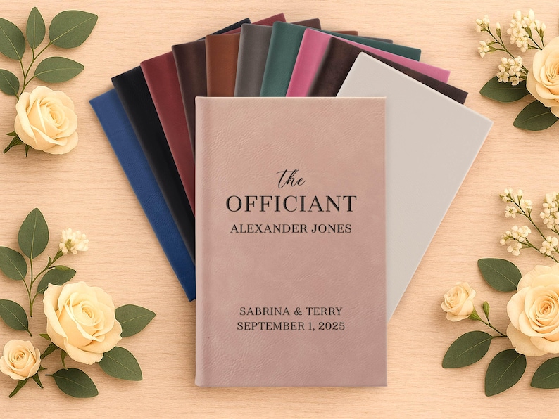 May include: A collection of personalized wedding officiant books in various colors, including pink, blue, and brown. The cover of the featured book is pink and reads "The Officiant" with a name and wedding date. Floral accents surround the books.