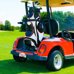 May include: A red golf cart with a black and white golf bag in the back. The golf cart has a license plate that says "Jennifer".