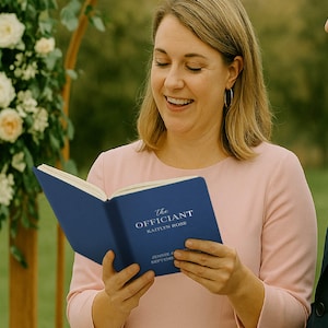 May include: A woman in a pink dress holds a blue book titled "The Officiant" with the name "Kaitlyn Ross" visible. The book is open, and she is smiling. A man in a navy suit stands to her right. A floral arch is in the background.
