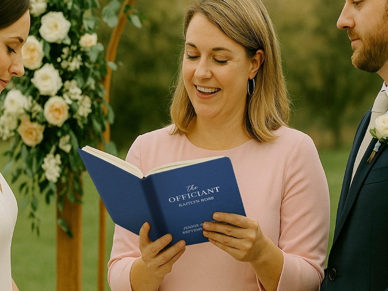 May include: A woman in a pink dress holds a blue book titled "The Officiant" with the name "Kaitlyn Ross" visible. The book is open, and she is smiling. A man in a navy suit stands to her right. A floral arch is in the background.