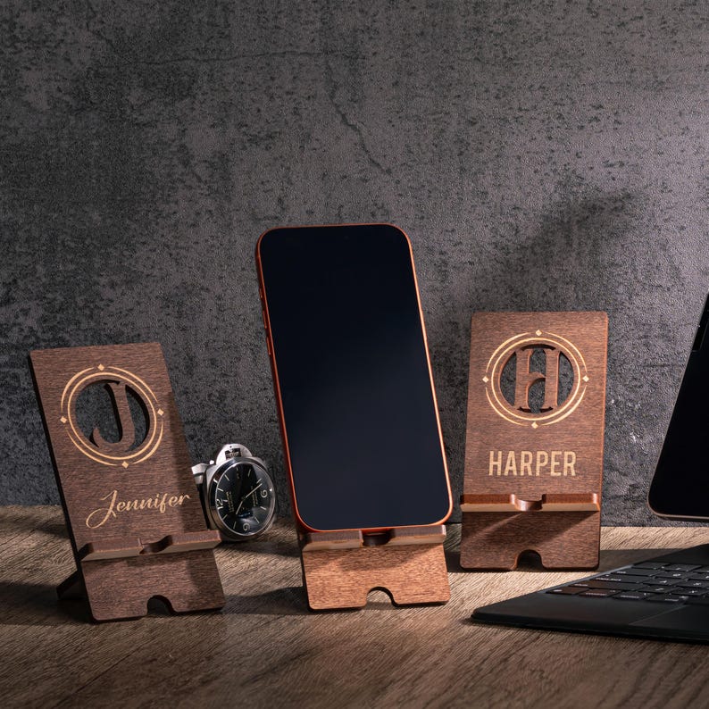 May include: Wooden phone stands with personalized initials and names. One stand displays the name "Jennifer", another features the initial "H" and the name "Harper". A smartphone is displayed on a stand. The stands are brown.
