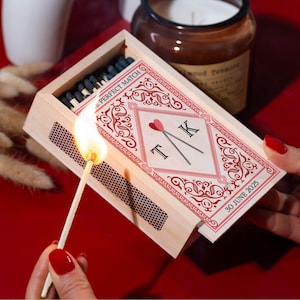 May include: A lit match ignites from a wooden matchbox with a decorative red and white design. The matchbox is labeled "PERFECT MATCH" and features initials "T K" with a heart. A candle and dried flowers are in the background.