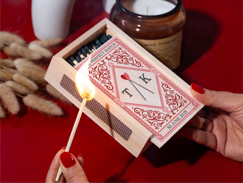 May include: A lit match ignites from a wooden matchbox with a decorative red and white design. The matchbox is labeled "PERFECT MATCH" and features initials "T K" with a heart. A candle and dried flowers are in the background.