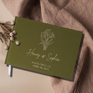 Op de afbeelding: Een groen gastenboek met een wit bloemenontwerp en de tekst "Henry & Sophia" en "Santa Cruz, CA April 08, 2021".