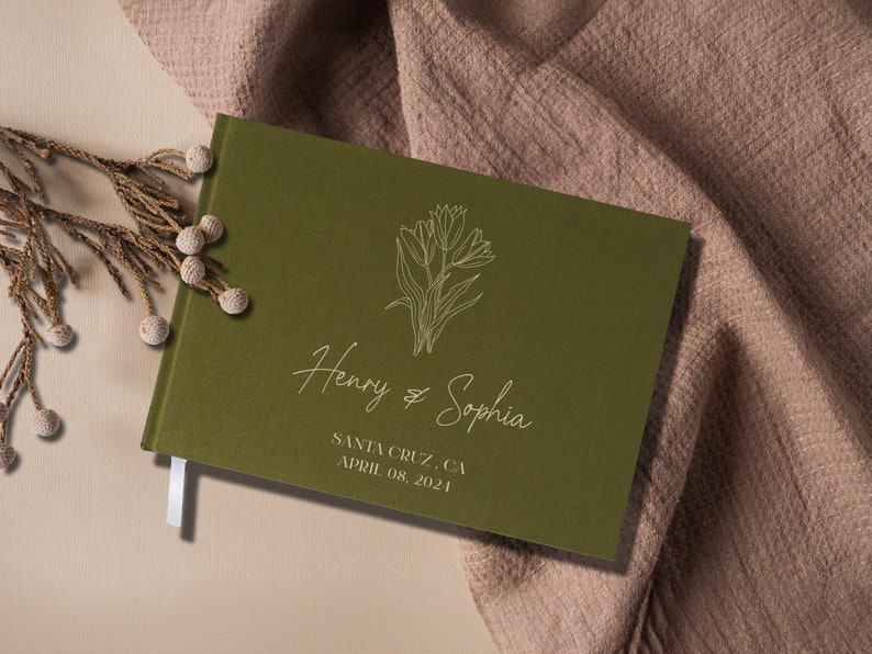 Op de afbeelding: Een groen gastenboek met een wit bloemenontwerp en de tekst "Henry & Sophia" en "Santa Cruz, CA April 08, 2021".