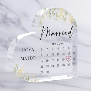 Puede incluir: Un recuerdo de acr&iacute;lico transparente en forma de coraz&oacute;n con un calendario que muestra la fecha 20 de junio de 2025, rodeado de un coraz&oacute;n rojo. Los nombres "NOVA + MATEO" est&aacute;n impresos debajo del calendario, y la palabra "Casado" est&aacute; impresa encima del calendario con un dise&ntilde;o floral.