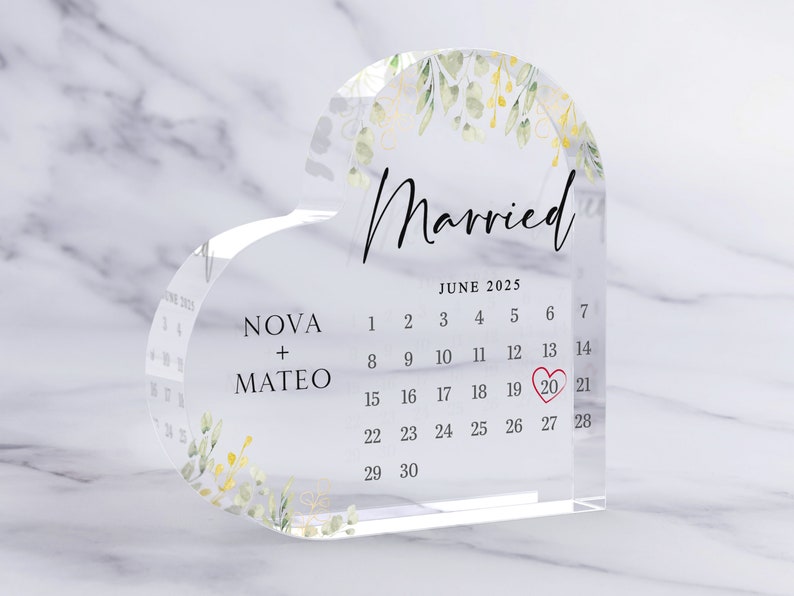 Puede incluir: Un recuerdo de acr&iacute;lico transparente en forma de coraz&oacute;n con un calendario que muestra la fecha 20 de junio de 2025, rodeado de un coraz&oacute;n rojo. Los nombres "NOVA + MATEO" est&aacute;n impresos debajo del calendario, y la palabra "Casado" est&aacute; impresa encima del calendario con un dise&ntilde;o floral.