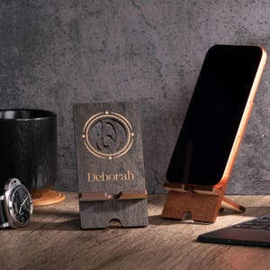 May include: A wooden phone stand with the name "Deborah" engraved on it, along with a decorative design. A smartphone is propped up on a wooden stand. A black watch and a black mug are also visible.