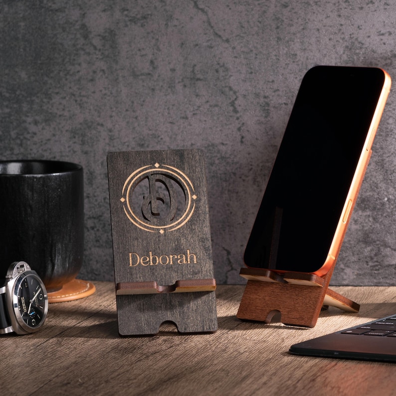May include: A wooden phone stand with the name "Deborah" engraved on it, along with a decorative design. A smartphone is propped up on a wooden stand. A black watch and a black mug are also visible.